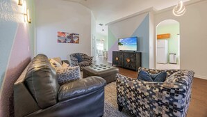 Un salon moderne avec un canapé, un pouf et un fauteuil à motifs. Un téléviseur à écran plat est fixé au mur, et on aperçoit un meuble en arrière-plan.