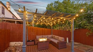 A covered outdoor seating area with a wooden deck, a cushioned bench, and a table with a light fixture.