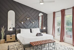 Quarto com cama, mesas de cabeceira, um banquinho, espelho e janela com cortinas.