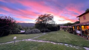 Paisaje rural con un atardecer, un árbol grande y un edificio con terraza.