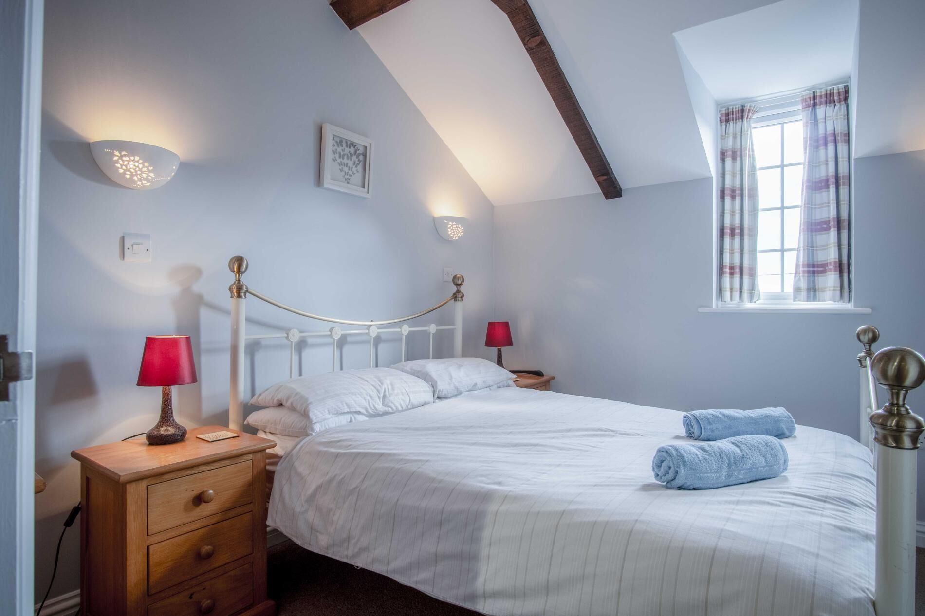 A bedroom with a bed, bedside lamps, a wooden nightstand, a window with curtains, and a picture on the wall.