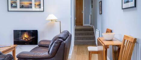 Een gezellige woonkamer met een leren bank, een open haard, een eettafel met stoelen, en een trap naar een andere kamer.