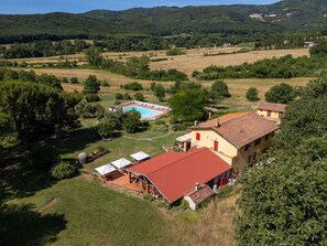 Una propietat rural amb piscina, una casa gran amb teulada vermella i vegetació al voltant.