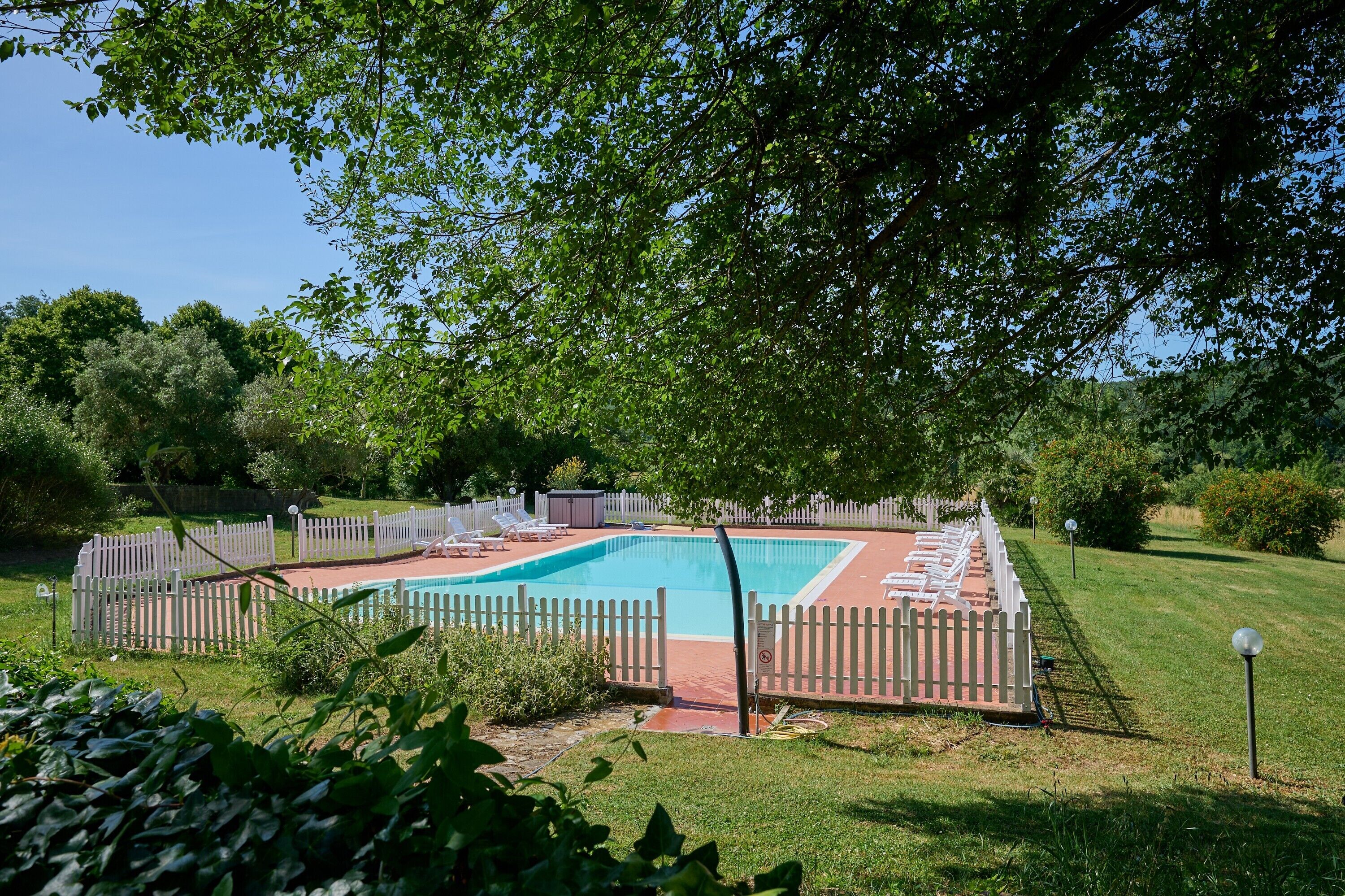 Ein rechteckiger Swimmingpool, umgeben von einem weißen Zaun, mit Liegestühlen und einem kleinen Gartenhaus im Hintergrund.