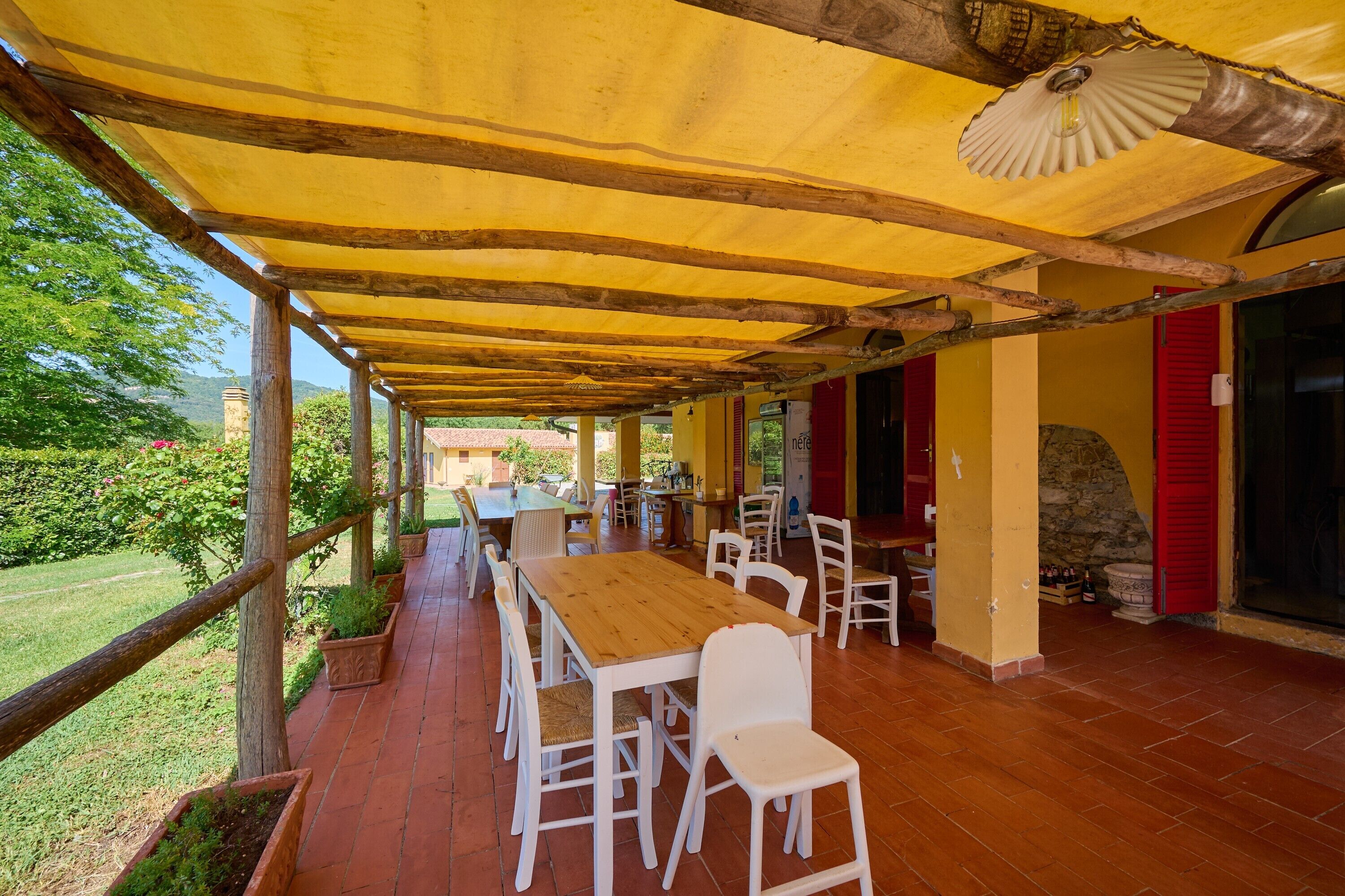 Une terrasse extérieure avec des tables en bois et des chaises blanches, un auvent jaune et un mur en pierre.