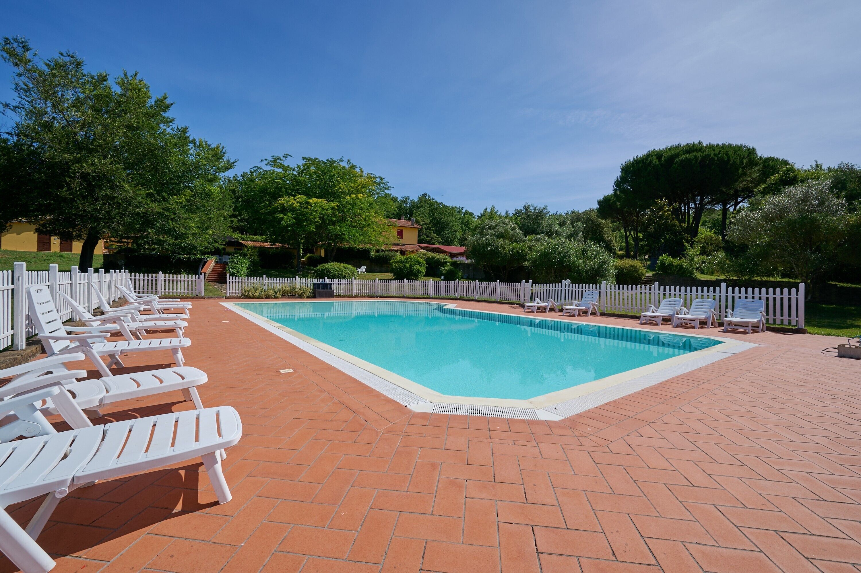 Une piscine rectangulaire entourée de chaises longues blanches, une clôture blanche en bois et une terrasse en briques.