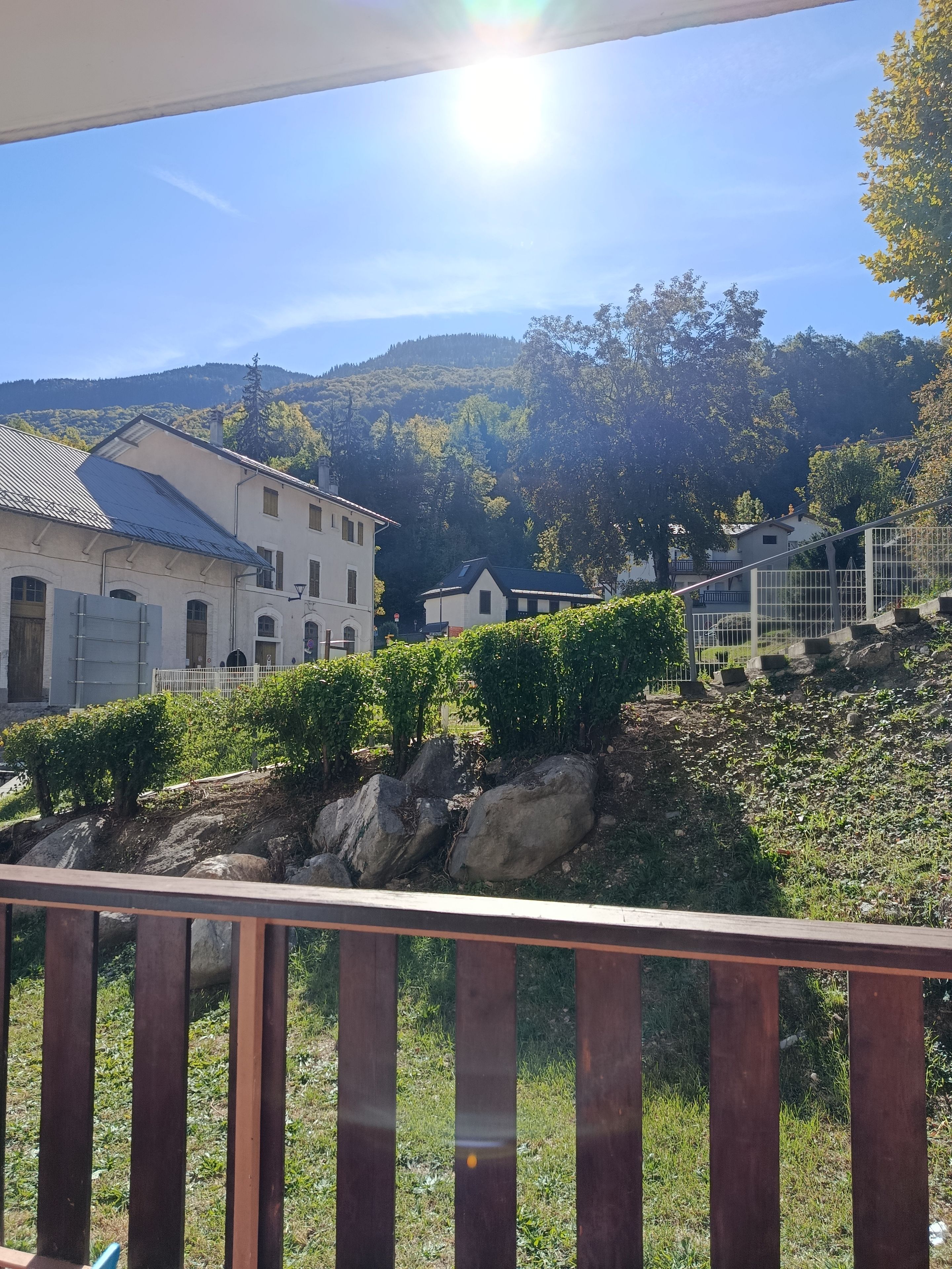 A view of a building with a sloped roof, a balcony with a wooden railing, and a hillside with greenery and rocks.