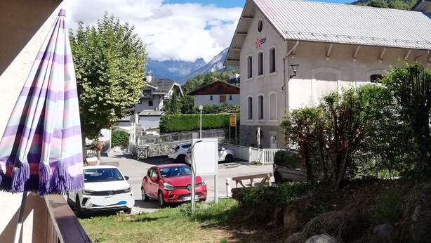 Un balcón con un paraguas a rayas, vista de coches aparcados, un edificio con un letrero y una montaña al fondo.
