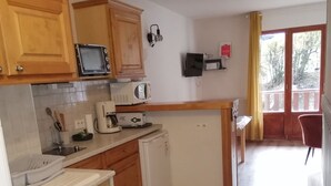 Cocina compacta con armarios de madera, microondas, tostadora y cafetera. Hay un lavamanos pequeño y un refrigerador. La cocina da a un balcón con vista a árboles.