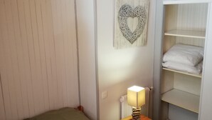 Un dormitorio pequeño y acogedor con dos camas individuales, una mesita de noche con lámpara, cabecero de madera, radiador y un armario blanco con estantes.