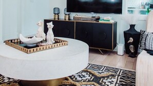 A modern living room with a large flat-screen TV, a round coffee table, and a patterned rug. - Discover Ponce Hideaway—an elegant 2BR, 2.5BA retreat in Midtown Atlanta. Modern-eclectic design, gold accents, and jewel tones set the scene. Enjoy a private patio, full kitchen, washer/dryer, & walkable access to Ponce City Market and Piedmont Park (Atlanta)