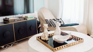 A modern living room with a large flat-screen TV, a black and gold cabinet, a round coffee table with decorative items, and a patterned rug. - Discover Ponce Hideaway—an elegant 2BR, 2.5BA retreat in Midtown Atlanta. Modern-eclectic design, gold accents, and jewel tones set the scene. Enjoy a private patio, full kitchen, washer/dryer, & walkable access to Ponce City Market and Piedmont Park (Atlanta)