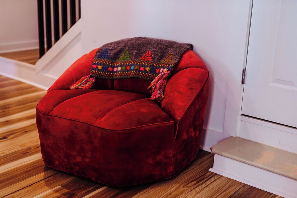 A red velvet armchair with a colorful throw blanket, positioned against a white wall with a wooden floor and a staircase in the background.