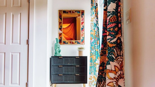 Un couloir avec une porte blanche, une commode noire à tiroirs, un miroir avec un cadre géométrique et un rideau à motifs.
