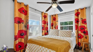 Quarto com cama, roupa de cama xadrez, ventilador de teto e janelas amplas com cortinas.