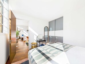 Une chambre d’hôtel moderne avec un plancher en bois, un lit recouvert d’une couverture à carreaux, un grand miroir et une vue sur la salle à manger adjacente.