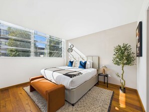 Une chambre moderne avec un grand lit, un banc, une plante en pot et une vue sur les bâtiments à travers la fenêtre.