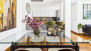 Una cocina moderna con gabinetes blancos, un comedor con mesa de vidrio y dos sillas, y un cuadro abstracto grande en la pared.