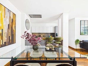 Una cocina moderna con gabinetes blancos, un comedor con mesa de vidrio y dos sillas, y un cuadro abstracto grande en la pared.