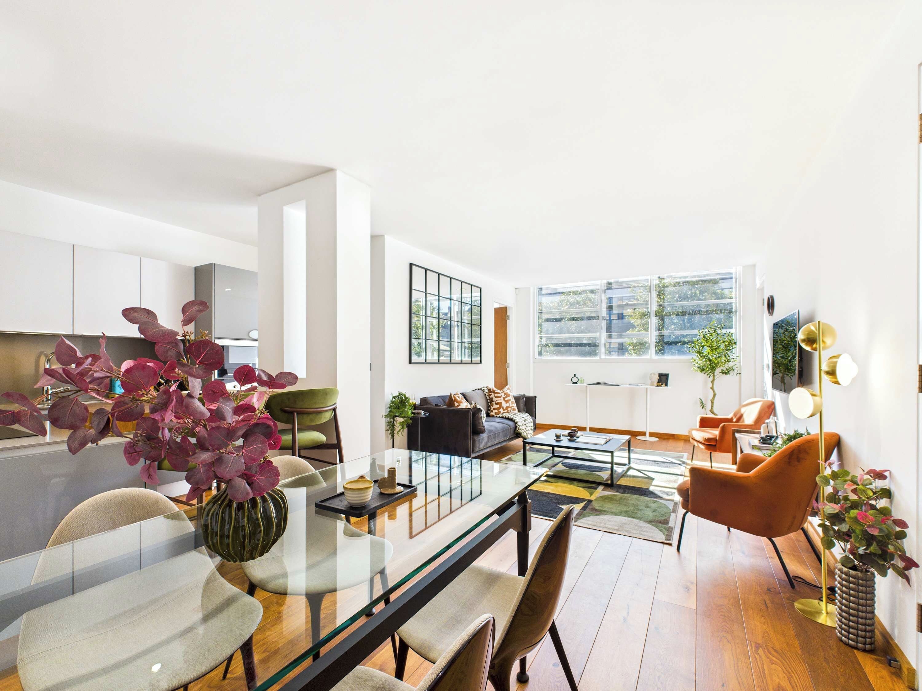 A modern living room with a glass dining table, a sofa, and a coffee table. There is a kitchen area with cabinets and a sink.