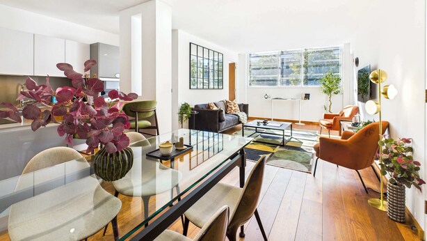 Una sala moderna con una mesa de comedor de vidrio, un sofá y una mesa de centro. Hay una zona de cocina con armarios y un lavamanos.