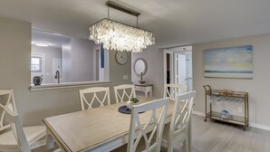 A dining area with a wooden table, white chairs, a chandelier, a wall clock, a mirror, and a painting. - Courtside 52- ground level 2BR flat! (Hilton Head Island)