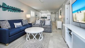 Un salon moderne avec un canapé bleu, des fauteuils à motifs, une table basse blanche et un téléviseur à écran plat diffusant une scène de plage.