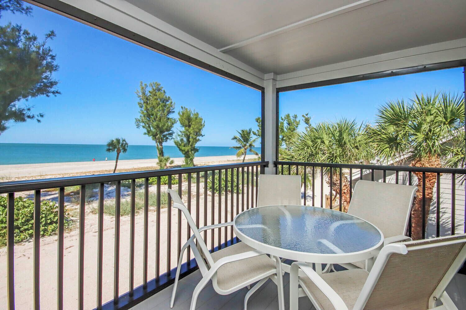 A balcony with a glass table and chairs, overlooking a beach with palm trees and the ocean.