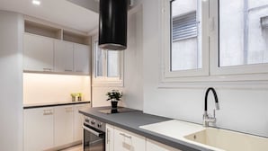Une cuisine moderne avec des meubles de couleur claire, une hotte noire, un four encastré et une grande fenêtre avec des stores.