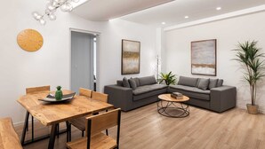 Un salon moderne avec une table à manger en bois, des canapés gris et une table basse. Deux tableaux abstraits ornent les murs, et une plante en pot est posée dans un coin.