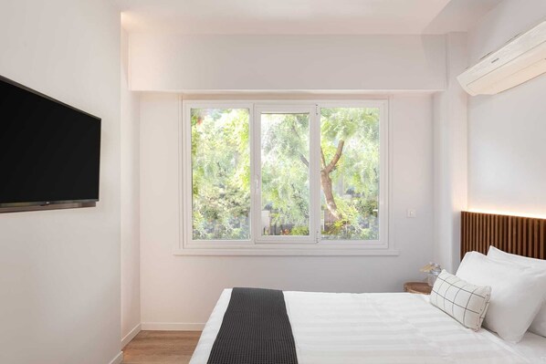 A bedroom with a bed, a TV mounted on the wall, a window with a view of trees, and a bedside table with a lamp. - Acropolis Holmes Residence, Apartment (Athens)
