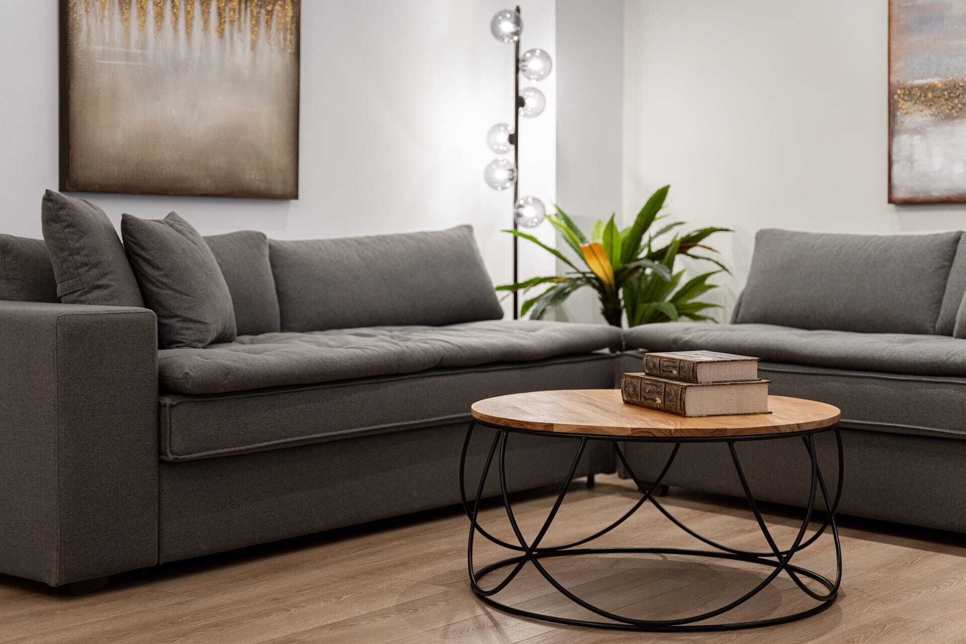 A modern living room with a grey sectional sofa, a round wooden coffee table with metal legs, and a plant in the corner.