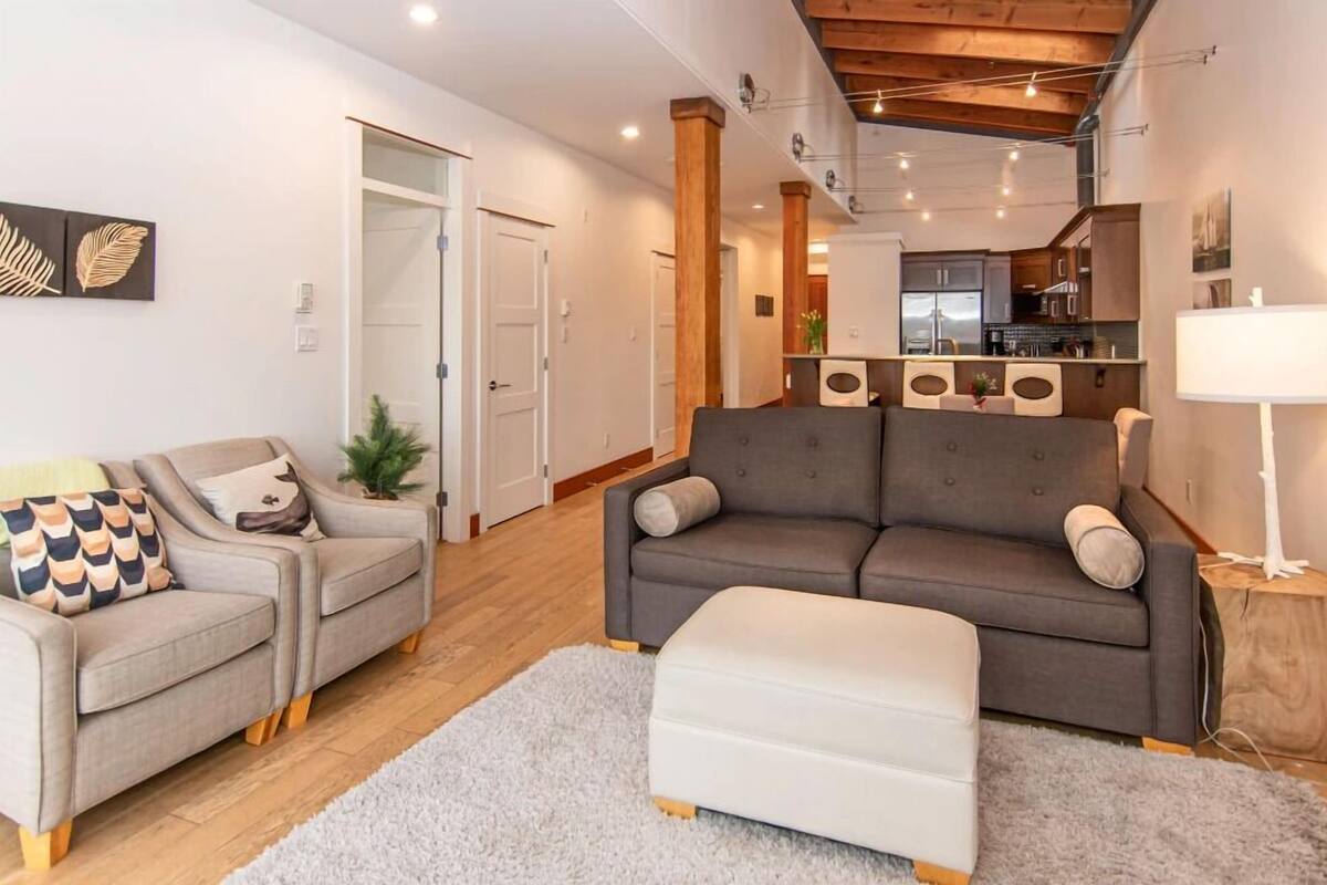 A modern living room with a grey sofa, a grey armchair, and a white ottoman. There is a kitchen in the background with wooden cabinets and a white countertop.