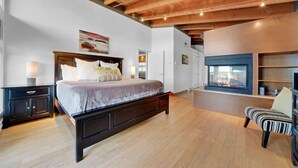 A bedroom with a large bed, wooden headboard, bedside tables, a chair, and a fireplace. - Mariner's Loft (Salt Spring Island)