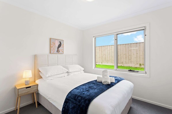Une chambre à coucher avec un lit, une table de chevet avec une lampe, une fenêtre donnant sur une clôture en bois et de l’herbe, et un tableau encadré accroché au mur.