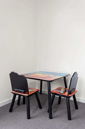 Une petite table carrée, dont le plateau présente un motif abstrait et coloré, est entourée de deux chaises noires, avec des assises et des dossiers orange.
