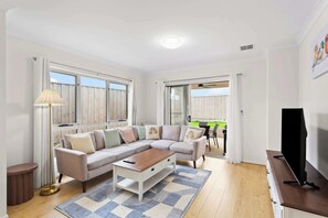 A living room with a beige sectional sofa, a coffee table, a TV stand, and a floor lamp. There is a sliding glass door leading to a balcony with a table and chairs. - Family-Friendly Coastal Retreat with Modern Amenities (Cowes)