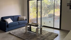 A modern living room with a blue sofa, a glass coffee table, and a view through large sliding glass doors to an outdoor area.