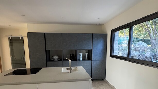 A modern kitchen with a large island, dark cabinets, and a window with a view of trees.