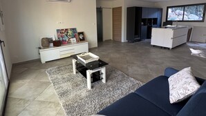 A modern living room with a blue sofa, a coffee table, and a rug. There is a kitchen area with cabinets and a window.