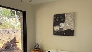 A bedroom with a bed, a wall-mounted artwork, a small table with a lamp, and a view of the outdoors.