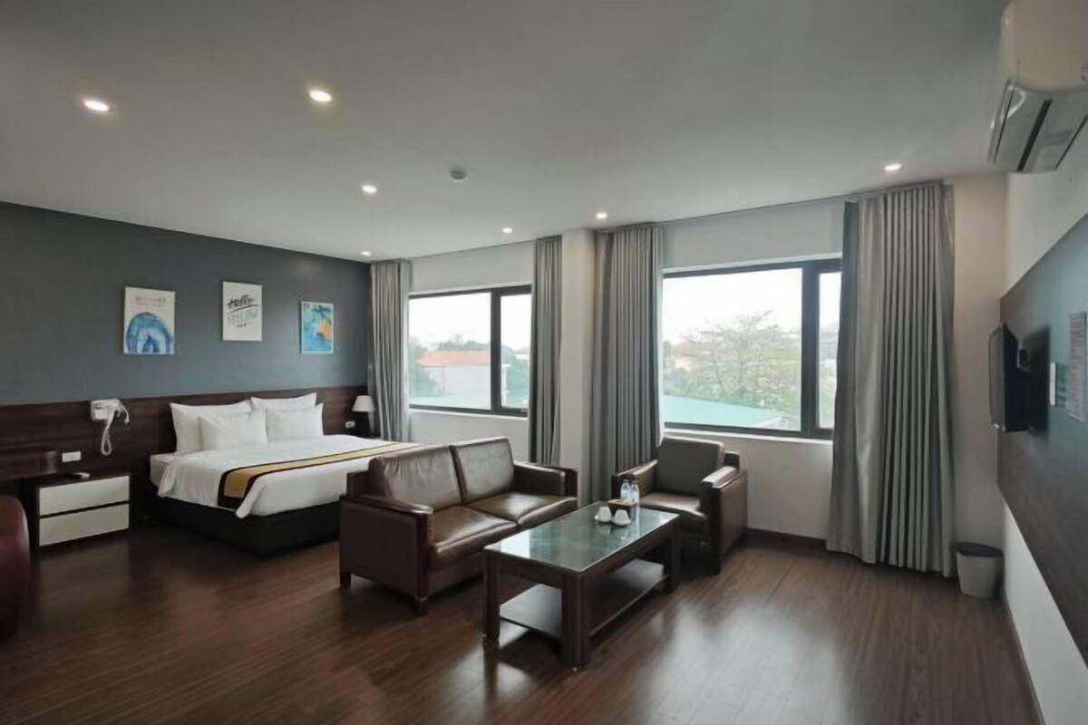 Une chambre d’hôtel avec un lit, deux canapés, une table basse, une télévision et de grandes fenêtres avec des rideaux.