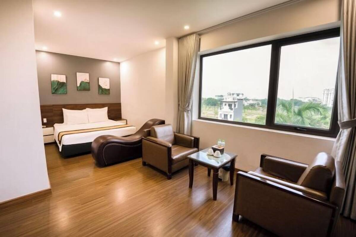 Une chambre d’hôtel avec un lit, deux fauteuils en cuir, une petite table et une grande fenêtre donnant sur des immeubles et des espaces verts.