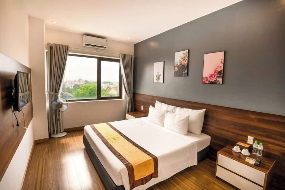 Une chambre d’hôtel avec un grand lit, une table de chevet équipée de divers accessoires, un téléviseur fixé au mur et une fenêtre avec des rideaux.