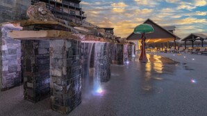 Une fontaine en pierre avec de l’eau en cascade, une grande structure en forme de parasol et un bâtiment avec des balcons en arrière-plan.