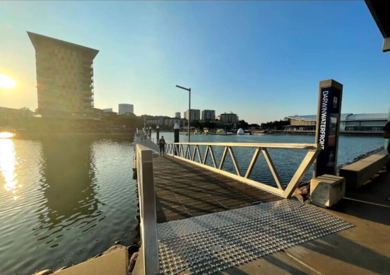 A pier with a metal railing, a sign reading "DARWIN WATERFRONT," and a city skyline in the background.