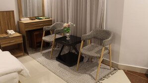 A hotel room with a bed, a wooden nightstand with a phone, a desk with a mirror, two grey upholstered chairs, a small black table with a vase of flowers, and a grey carpet. - Rafa Serviced Apartments - Economic (Riyadh)