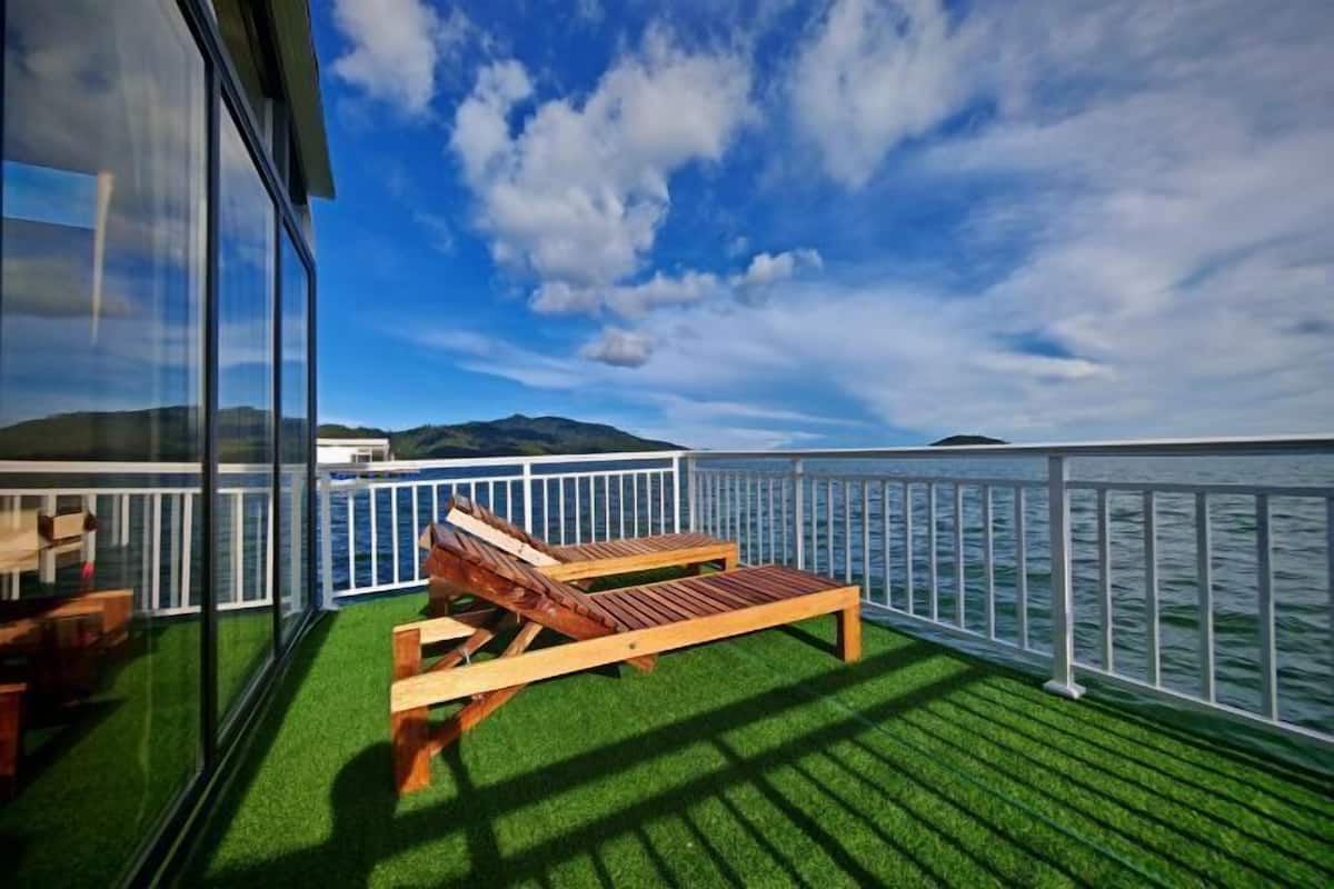 Eine Terrasse mit künstlichem Rasen, einem Holzliegenstuhl und einem weißen Geländer mit Blick auf ein Gewässer.