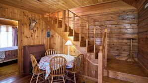 A wooden cabin interior with a dining table, chairs, a lamp, a staircase, and a deer head mounted on the wall.