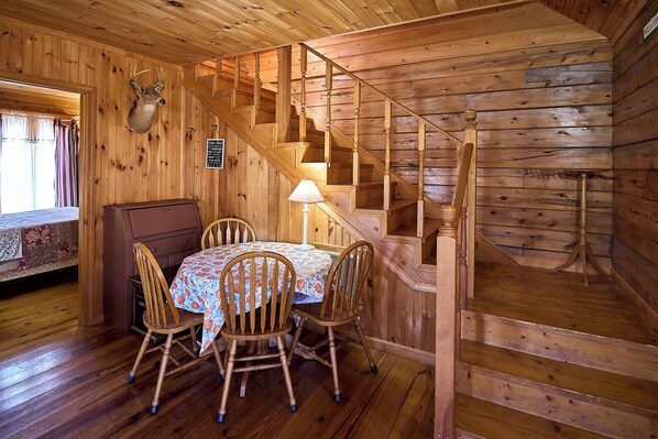 A wooden cabin interior with a dining table, chairs, a lamp, a staircase, and a deer head mounted on the wall.